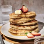 A stack of Cinnamon Buttermilk Pancakes topped with a pat of butter and sliced strawberries sits on a white plate, with a glass of water and a jar of jam nearby.
