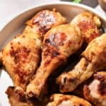 A plate of cooked, browned Chipotle Mango Chicken drumsticks with a golden, slightly crispy skin. A green leafy garnish and other dishes are blurred in the background.