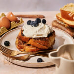 French Toast with Vanilla Bean Whipped Cream topped with fresh blueberries on a plate, accompanied by a fork and knife, with eggshells and slices of bread visible in the background.