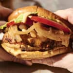 Hands holding a Juicy Chicken Burger with lettuce, tomato, sautéed onions, and a creamy sauce on a toasted bun, over a plate.