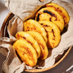 A woven basket lined with a beige cloth holds six golden, grilled arepas, arranged neatly and overlapping each other—a tempting display that highlights the delicious tradition of arepas.