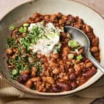 A bowl of Pork and Beef Chili with beans and ground meat, topped with sour cream, chopped green onions, and herbs. A spoon rests inside the bowl, which sits on a textured beige cloth.