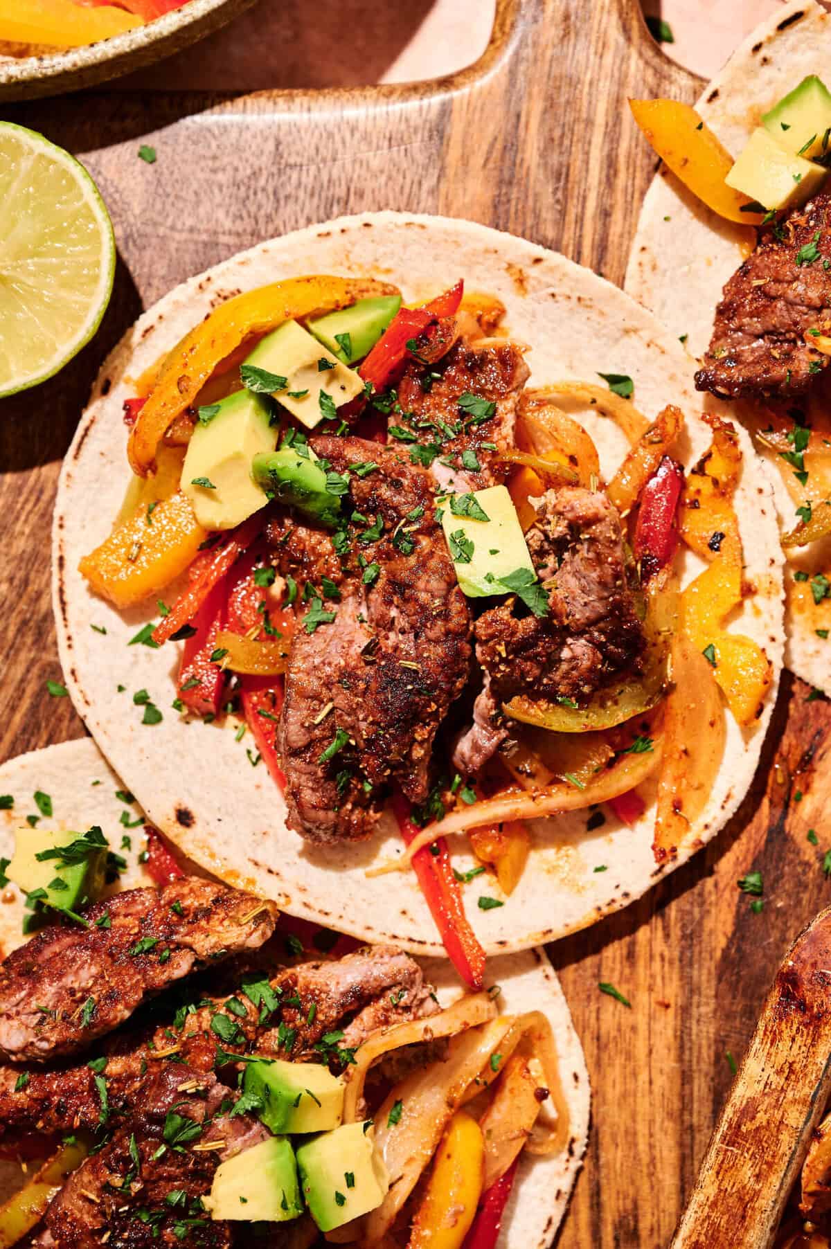 Three steak fajitas on tortillas with grilled bell peppers, onions, chopped avocado, and cilantro on a wooden board. A lime half is visible in the corner, making these 15-Minute Sheet Pan Fajitas as vibrant and fresh as they are quick to prepare.