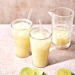 Two glasses of frothy Brazilian Limeade with straws are on a pink surface next to a glass pitcher filled with more of the refreshing drink. Sliced and whole limes are scattered nearby, highlighting the fresh, zesty flavors of this vibrant beverage.