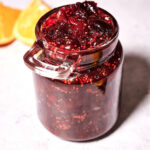 A glass jar filled with chunky dark red berry jam, reminiscent of Homemade Cranberry Sauce, sits on a light surface, with orange slices blurred in the background.