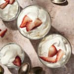 Three glasses of homemade whipped cream topped with sliced strawberries, surrounded by several spoons and a small bowl of extra strawberry slices on a textured surface.