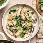 A plate of creamy Mushroom and Sun Dried Tomato Orzo with spinach, topped with fresh herbs, invites you to dig in. A fork rests on the plate, while vibrant fresh ingredients surround the dish.