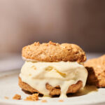 An ice cream sandwich with vanilla ice cream and a swirl of nut butter between two crumbly cookies sits on a white plate. Some ice cream is melting and oozing out, and there are crumbs scattered around.