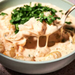 A bowl of creamy Poblano Rajas Gnocchi topped with chopped fresh herbs. A spoon lifts a portion, showing the thick, cheesy sauce stretching. The bowl sits on a speckled surface.