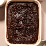 A rectangular baking dish filled with rich Sweet Potato Brownies, topped with chocolate chips, surrounded by two glasses of milk and a stack of silver spoons on small plates.