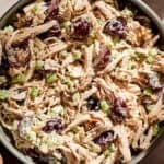 A bowl of Grape and Walnut Chicken Salad features shredded chicken, sliced grapes, chopped celery, and walnuts, all mixed in a creamy dressing. Nearby are grapes, walnuts, and sliced bread on the table.