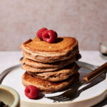 A stack of Chocolate Milk Pancakes topped with three raspberries sits on a white plate with a fork and a cinnamon stick. More raspberries are scattered nearby.
