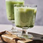 Two frosty green beverages with ice in clear glasses on a wooden board, with cinnamon sticks scattered nearby. The drinks appear creamy and have a subtle layered effect, perfectly capturing the unique blend of Oat Milk Matcha Horchata.