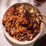 A plate of shredded Slow-cooked Crockpot Brisket with tender cooked onions and red peppers is served with gold utensils. A small pitcher of dark sauce sits beside the plate on a light surface.