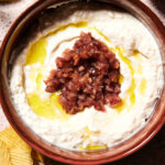 A ceramic bowl filled with creamy whipped sheep’s milk feta dip, topped with olive oil and a chunky red mixture, surrounded by wavy potato chips.