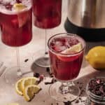 Three glasses of iced red hibiscus tea with lemon wedges, surrounded by lemon slices, dried hibiscus petals, and a jar of petals on a light counter—perfect for enjoying as an invigorating Hibiscus and Date Lemonade.