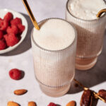 Two glasses of frothy Raspberry Almond Milk smoothie with gold straws, surrounded by a bowl of raspberries, loose raspberries, almonds, and dates, on a light surface.