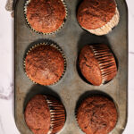 A muffin tin with six baked carrot muffins, three of which are tilted in their cups, on a marble countertop with silver utensils nearby.