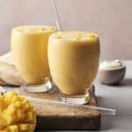 Two glasses of mango lassi with glass straws sit on a wooden board, evoking the refreshing appeal of Frozen Mango Lemonade. A diced mango and a small bowl of yogurt are nearby, all placed on a light tabletop with a pale background.