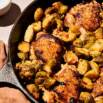 A cast iron skillet filled with Honey Dijon Chicken Thighs and Brussels sprouts, with a brown cloth underneath and a small dish in the background.