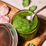 A spoon rests in a small jar filled with green pesto sauce on a wooden board, with fresh basil leaves and garlic cloves nearby—a scene that captures the essence of the Perfect Pesto Recipe.