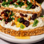 Close-up of seeded multigrain toast topped with whipped feta, dried cranberries, chopped almonds, fresh herbs, and a drizzle of honey, arranged on a white plate.