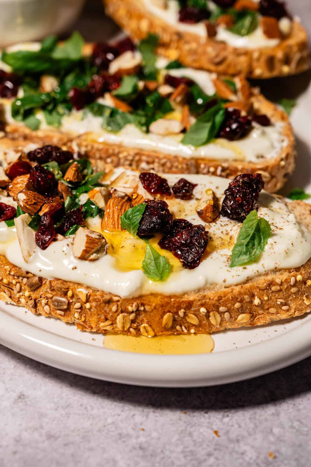 Close-up of seeded multigrain toast topped with whipped feta, dried cranberries, chopped almonds, fresh herbs, and a drizzle of honey, arranged on a white plate.