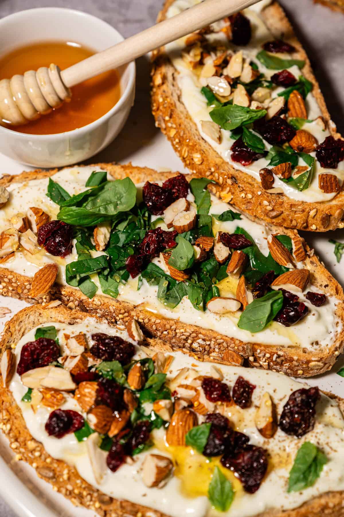 A platter of whipped feta toast topped with almonds, cranberries, fresh herbs, and honey, served with a small bowl of honey.