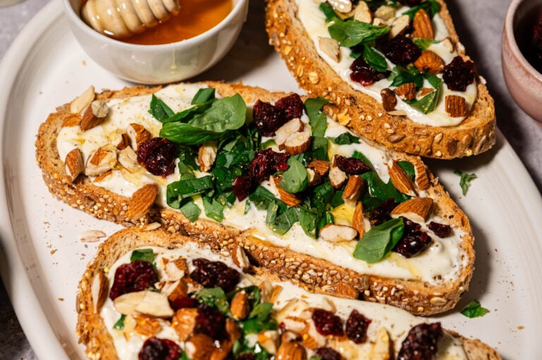 Toasted seeded sourdough slices topped with whipped feta, chopped fresh herbs, dried cranberries, and crushed almonds, drizzled lightly with honey, served on a white platter with a small bowl of honey on the side.