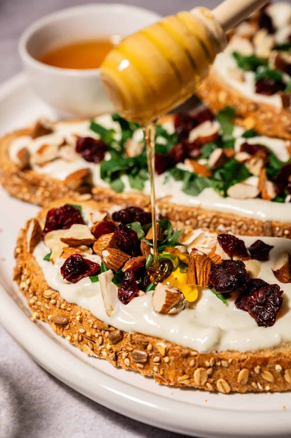 Three slices of whipped feta toast topped with chopped herbs, almonds, dried cranberries, and honey on a white platter.