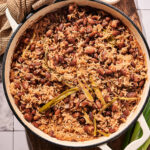 A pot of Coconut Rice and Beans with green onions sits on a wooden board, accompanied by a small bowl of seasoning and a textured kitchen towel nearby.