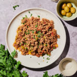A plate of Instant Pot Picadillo with ground meat, peppers, and herbs sits on a table with fresh parsley, a bowl of green olives, a pepper grinder, and a small bowl of salt.