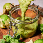 Roasted Tomatillo Salsa being poured into a glass jar, surrounded by fresh cilantro and tomatillos on a wooden surface.