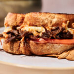 Close-up of a Birria Grilled Cheese sandwich filled with shredded beef, melted cheese, and a slice of tomato, served on a plate with a blurred bowl in the background.