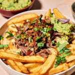 A bowl of crispy fries topped with shredded beef, guacamole, cheese, chopped green onions, red onion, and cilantro—these Loaded Birria Fries are bursting with flavor. A bowl of guacamole is in the background.