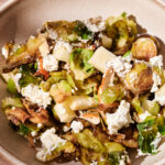 A bowl of Roasted Brussels Sprouts with Almond and Goat Cheese salad, featuring crumbled cheese, sliced almonds, and pieces of apple, served in a beige ceramic bowl on a light surface.