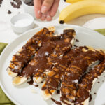 A hand sprinkles salt over sliced bananas drizzled with chocolate on a white plate, reminiscent of a delicious Peanut Butter and Chocolate Banana treat. Two whole bananas, a bowl of salt, and chocolate chips are on the table in the background.
