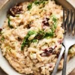 A bowl of Creamy Lemon Ricotta Orzo with sun-dried tomatoes, a green pesto drizzle, and cracked black pepper, served with two forks on the side.