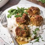 Four curry lamb meatballs are served on a bed of white rice, topped with sauce and garnished with chopped herbs. A fork rests on the plate, with extra herbs and a cutting board visible in the background.