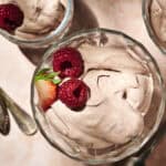 Glass bowls filled with chocolate mousse, each topped with raspberries and a strawberry slice, are finished with a generous dollop of Chocolate Protein Whipped Cream. Spoons are placed beside the bowls on a light-colored surface.