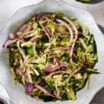 A vibrant Cucumber Spring Salad awaits in a bowl, featuring shredded zucchini, sliced red onions, green peas, and spices, all beautifully garnished with red pepper flakes. Lime wedges and small bowls of spices and honey rest nearby on the light-colored surface.