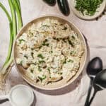 A bowl of creamy Jalapeño Popper Potato Salad garnished with chopped herbs, surrounded by green onions, jalapeños, a small bowl of herbs, a cup of milk, and two spoons on a napkin.