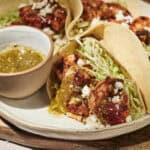 Three tacos filled with grilled chicken, shredded lettuce, and crumbled cheese are served on a plate with a small bowl of green salsa on the side—perfect for anyone who loves classic flavors or is looking to try something new like shrimp tacos.