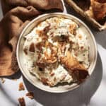A bowl of creamy Cannoli Dip, topped with broken cannoli shell pieces, sits on a light surface next to a brown napkin and a basket of whole cannoli shells.