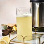 A glass pitcher of pale yellow ginger lemonade sits on a tray, with fresh lemon, ginger, and a stainless steel appliance in the background—a refreshing display of Ginger Lemon Infused Water waiting to be enjoyed.