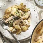 A plate of pasta shells mixed with grilled chicken, mushrooms, and herbs sits on a decorative white plate with two forks beside it, next to a partial view of a pot and a lemon, evoking the fresh flavors of Creamy Lemon Asparagus Pasta.