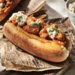 A toasted bun filled with seasoned shrimp, inspired by classic Langostino Rolls, is topped with creamy sauce and herbs, and sits on newspaper-style parchment atop a wooden board. Another sandwich and a dip are in the background.