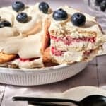 A No-bake Berry Icebox Cake, layered with whipped cream, strawberries, and blueberries on top, sits in a white dish. A slice is being lifted out. Two black spoons and small plates are nearby on the table.