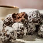 A stack of No-Bake Chocolate Protein Balls, coated in shredded coconut, with one truffle showing a bite taken out, sits on a light surface with a blurred cup in the background.