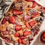 A Strawberry Almond French Toast Casserole baked to golden perfection is topped with sliced strawberries and powdered sugar in a rectangular dish, set next to a striped cloth and a glass of brown liquid.
